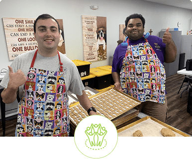 Two smiling men wearing colorful aprons, holding chocolate bars in a kitchen.