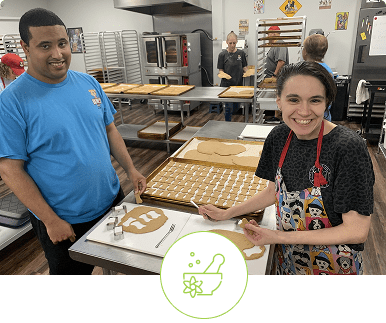Two people smiling and holding trays of baked goods in a kitchen.